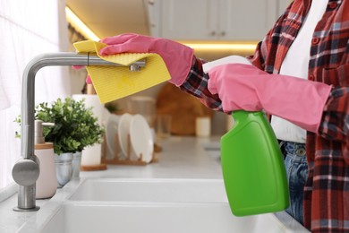 Photo of Woman with spray bottle and microfiber cloth cleaning water tap in kitchen, closeup