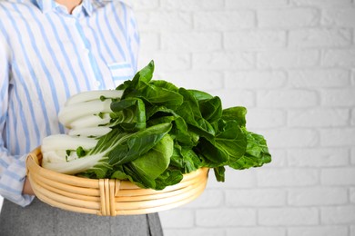 Photo of Woman holding wicker basket with bok choy cabbage near white brick wall, closeup