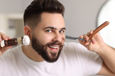 Photo of Handsome young man holding blade and shaving brush with foam in bathroom