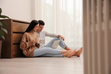 Photo of Happy couple with laptop sitting on warm floor at home. Heating system