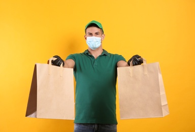 Photo of Courier in medical mask holding paper bags with takeaway food on yellow background. Delivery service during quarantine due to Covid-19 outbreak