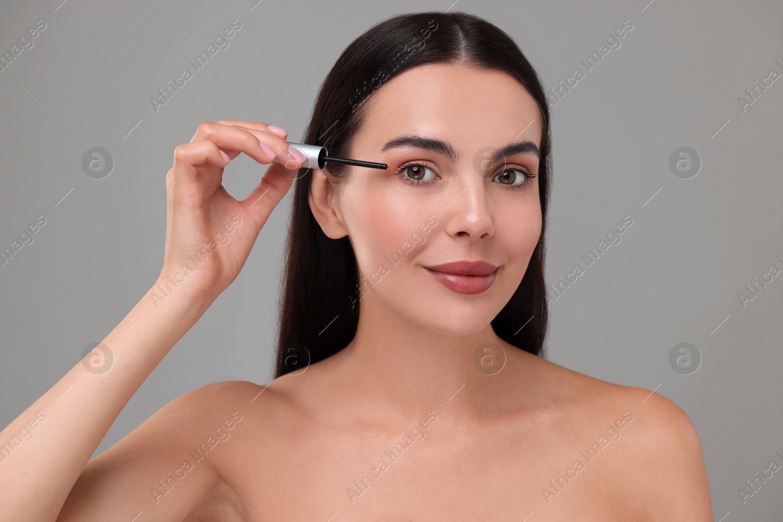 Photo of Beautiful woman applying serum onto her eyelashes on grey background. Cosmetic product
