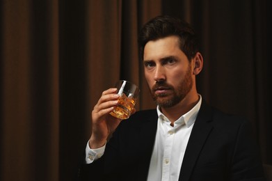 Photo of Man in suit holding glass of whiskey with ice cubes on brown background. Space for text