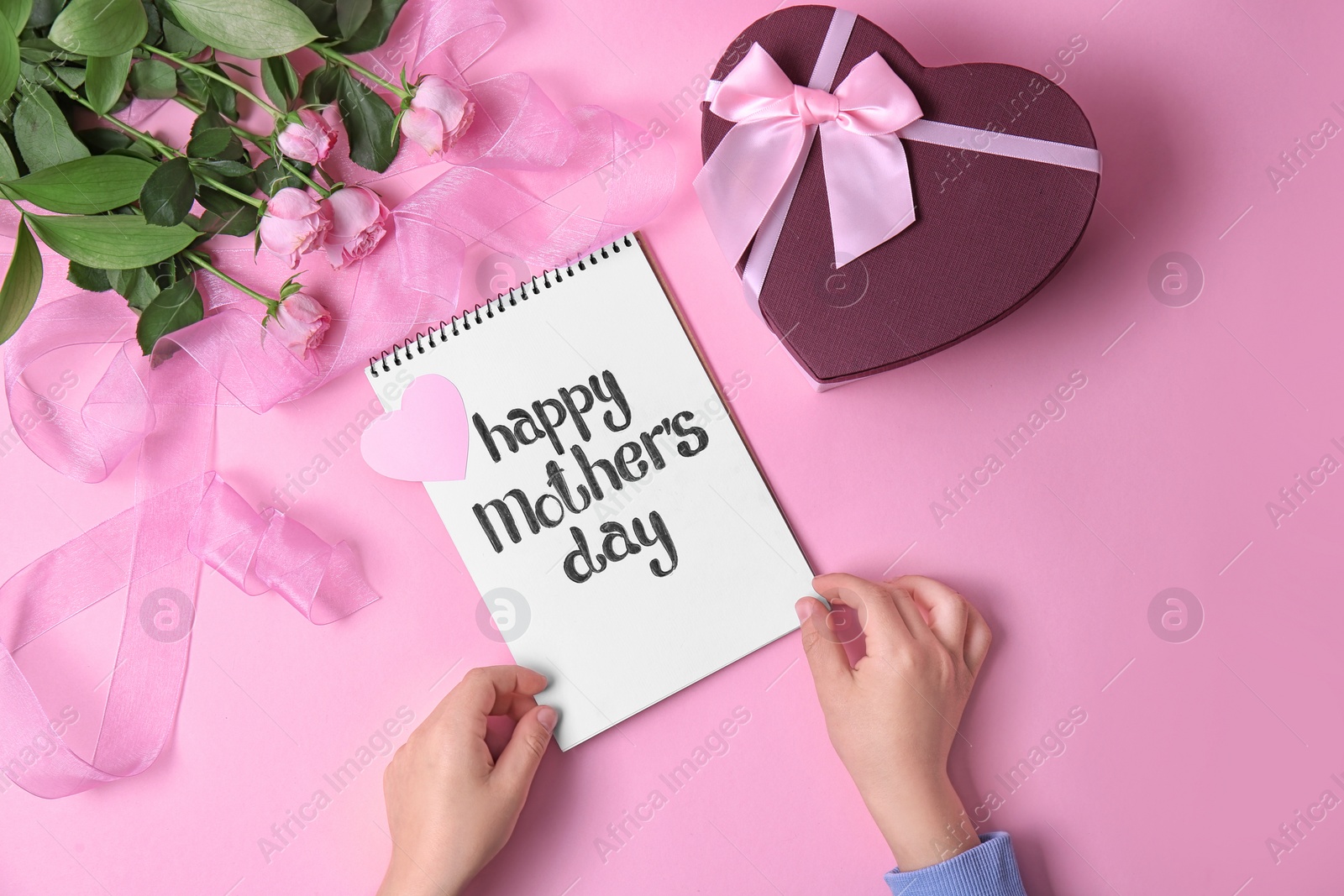 Photo of Child holding notebook with phrase "HAPPY MOTHER'S DAY" near gift and roses on table