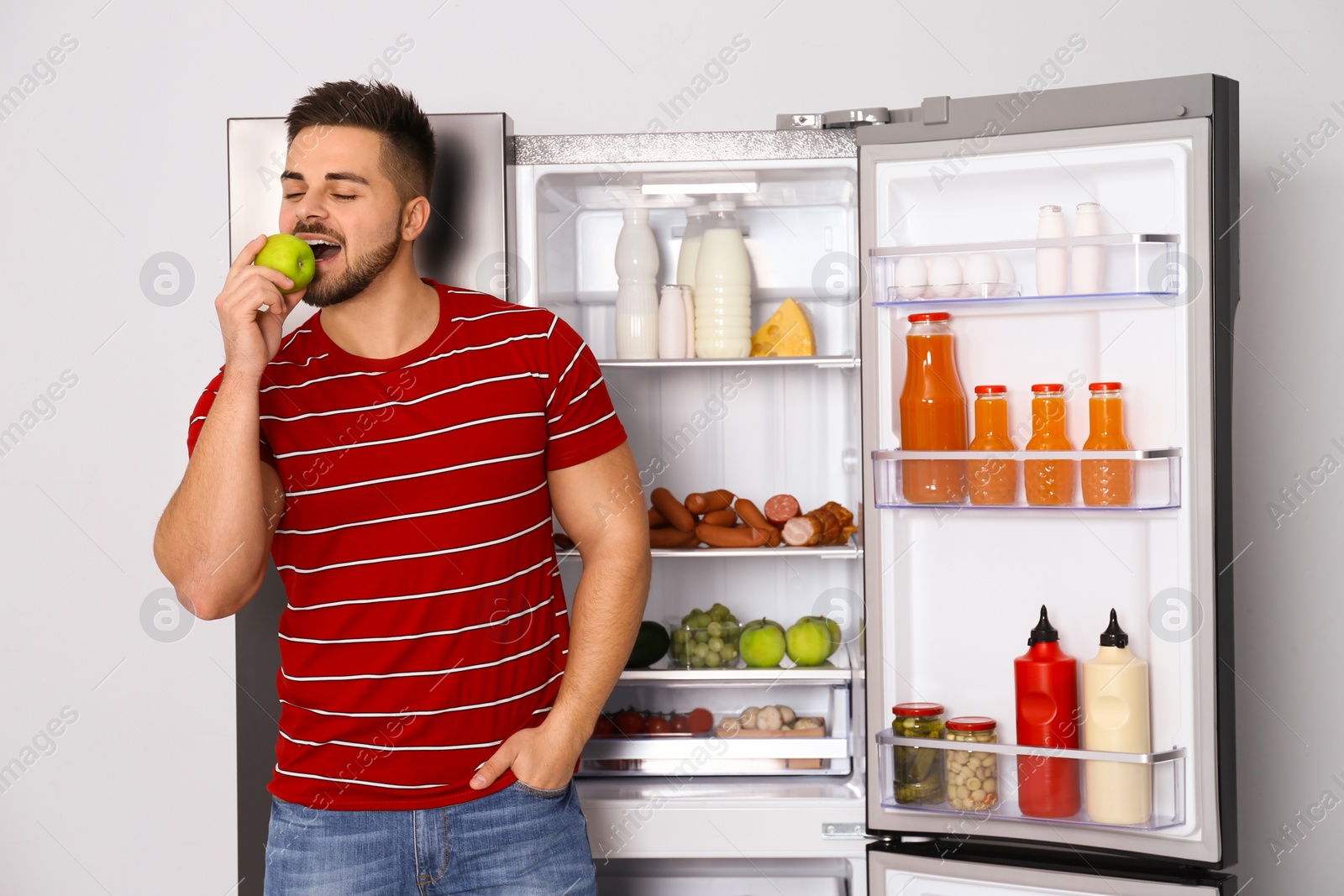 Photo of Happy young man eating apple near open refrigerator indoors