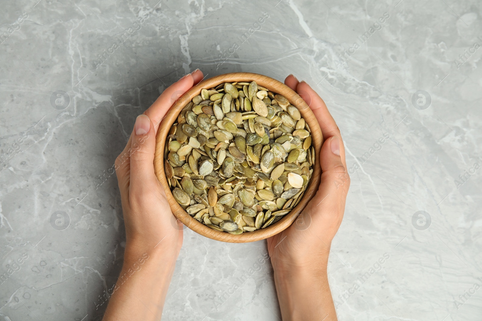 Photo of Young woman with bowl of raw pumpkin seeds at light grey marble table, top view