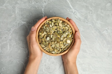 Photo of Young woman with bowl of raw pumpkin seeds at light grey marble table, top view