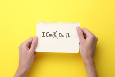 Photo of Motivation concept. Woman holding paper with changed phrase from I Can't Do It into I Can Do It by crossing over letter T on yellow background, top view