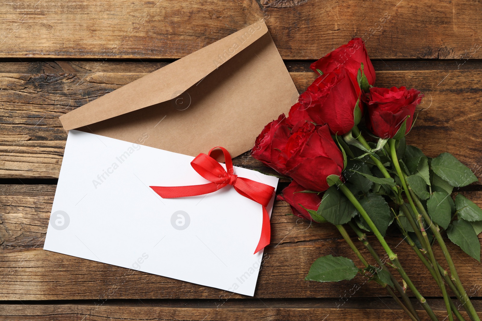 Photo of Blank greeting card, roses and envelope on wooden table, flat lay. Valentine's day celebration