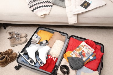 Open suitcase with warm clothes, accessories and shoes near sofa indoors