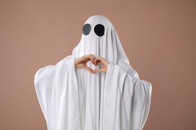 Photo of Cute ghost. Woman covered with white sheet making heart with hands on dark beige background