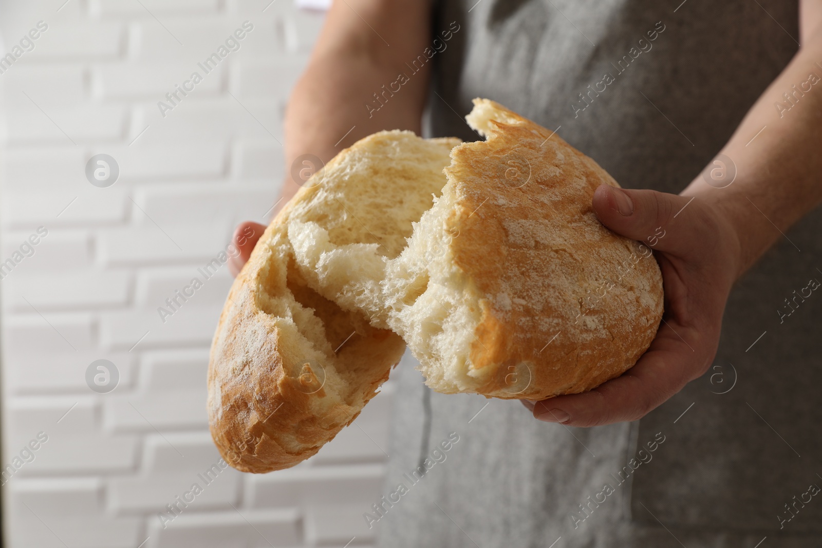 Photo of Man breaking loaf of fresh bread near white brick wall, closeup