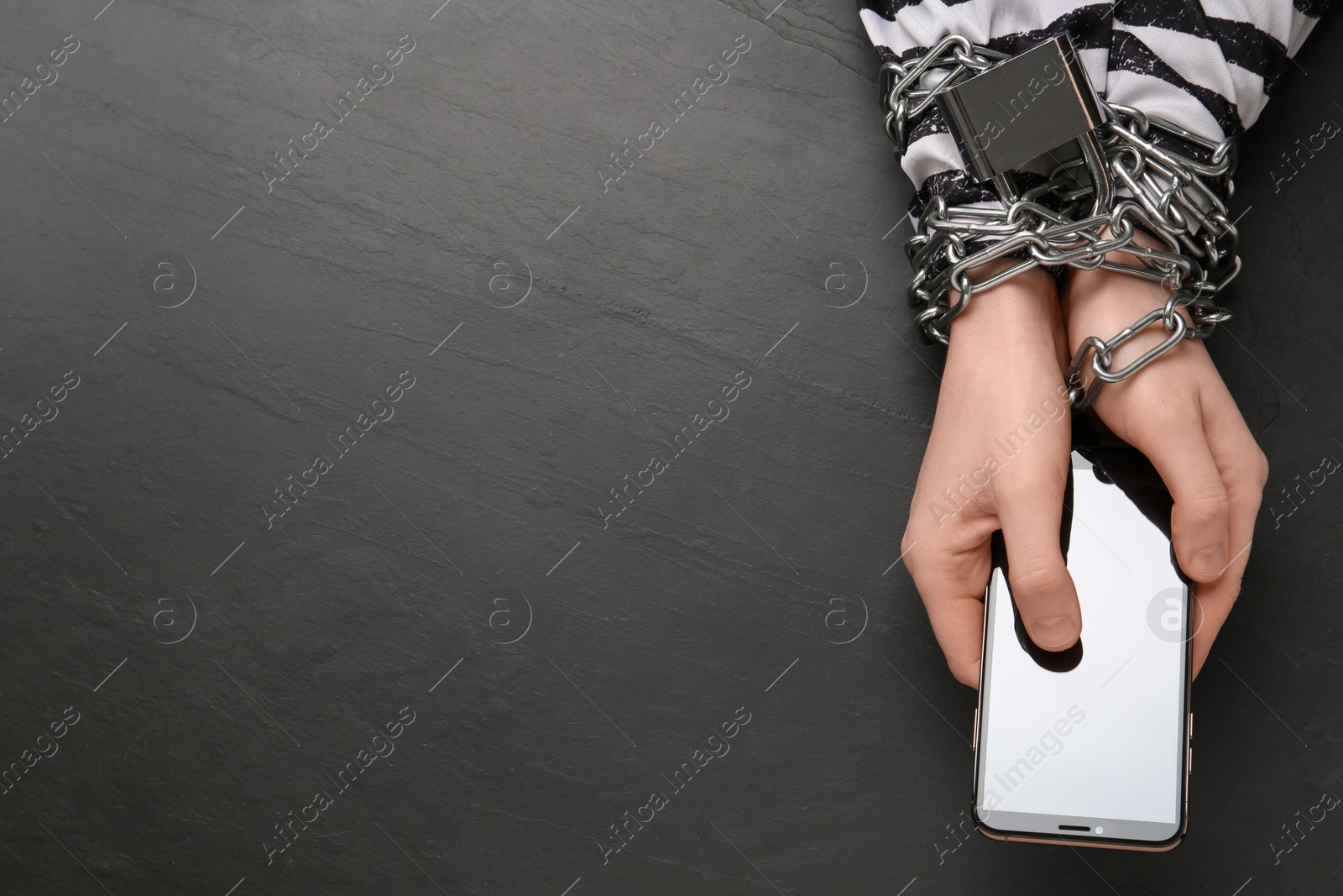 Photo of Prisoner holding smartphone in chained hands at black table, top view. Internet addiction