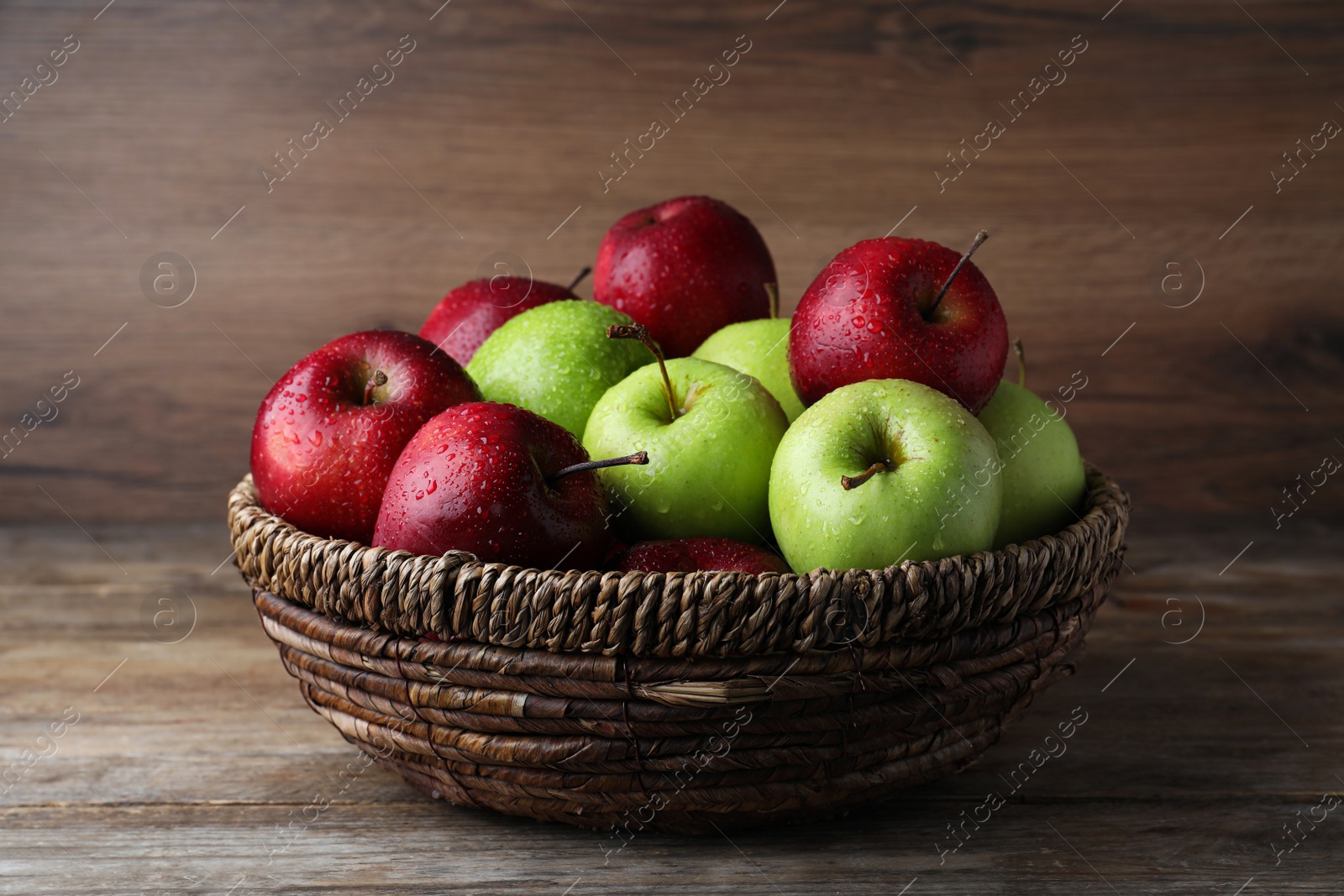 Photo of Fresh ripe green and red apples with water drops in wicker bowl on wooden table