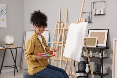 Photo of Young woman mixing paints on palette with brush near easel in studio