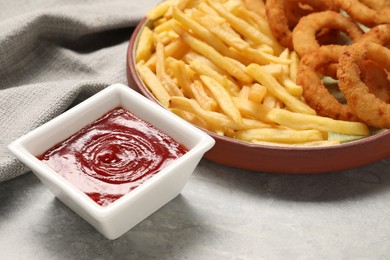 Tasty ketchup with fries and onion rings on light grey table, closeup