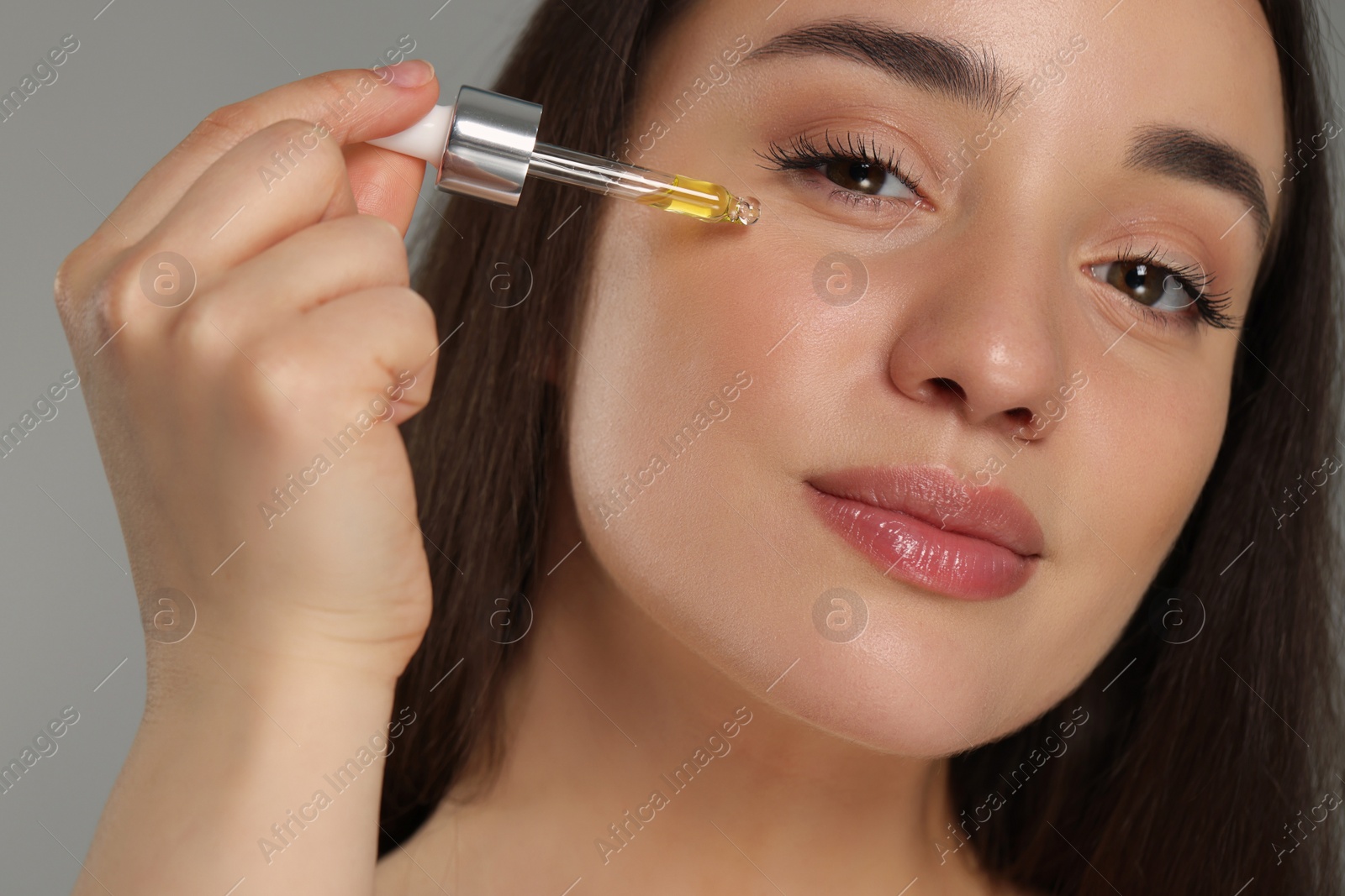 Photo of Beautiful young woman applying essential oil onto face on grey background, closeup