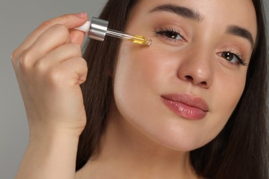 Beautiful young woman applying essential oil onto face on grey background, closeup