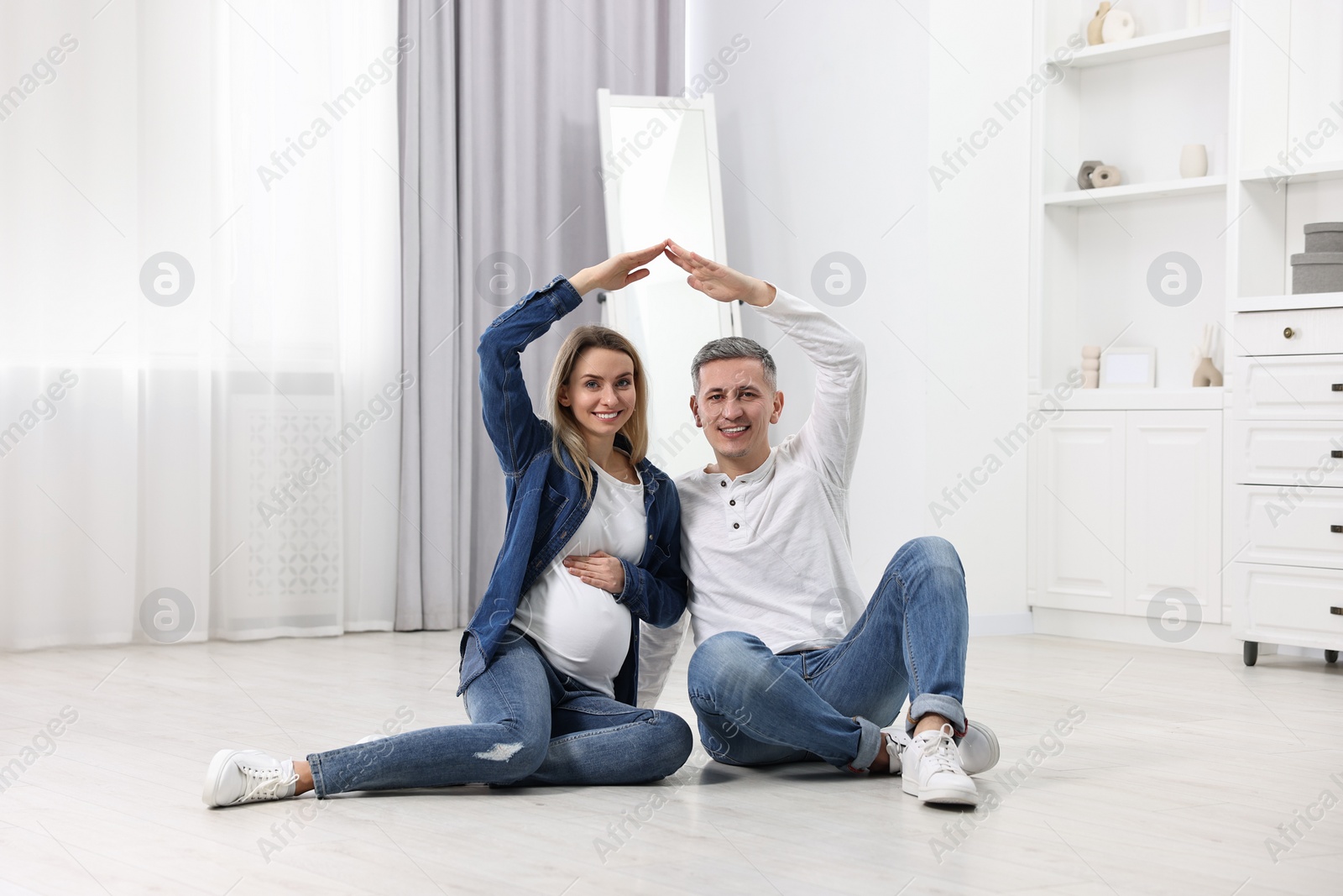 Photo of Young family housing concept. Pregnant woman with her husband forming roof with their hands while sitting on floor at home