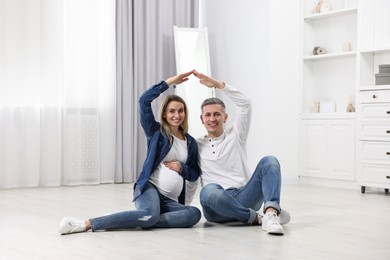 Young family housing concept. Pregnant woman with her husband forming roof with their hands while sitting on floor at home