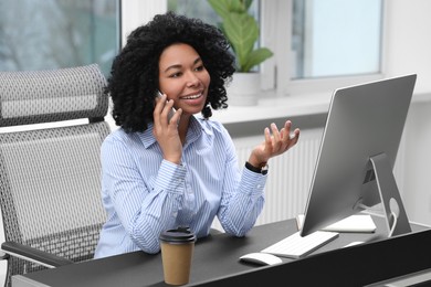 Photo of Young woman talking on phone while working with computer at table in office