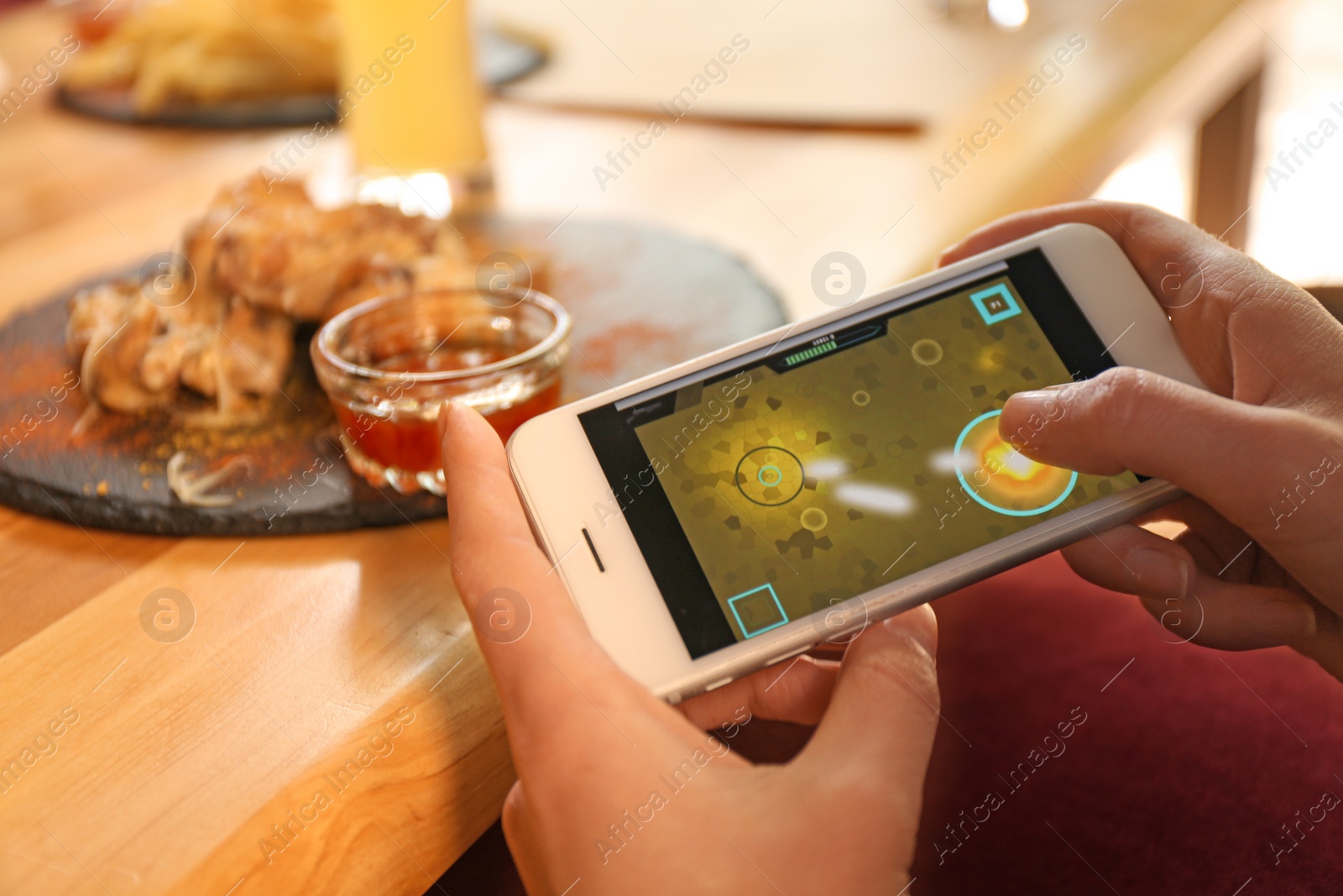 Photo of Woman playing game using smartphone at table with tasty BBQ wings in cafe, closeup