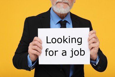 Photo of Unemployed senior man holding cardboard sign with phrase Looking For A Job on yellow background, closeup