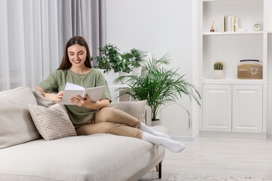 Photo of Beautiful young woman reading book on sofa in room with green houseplants