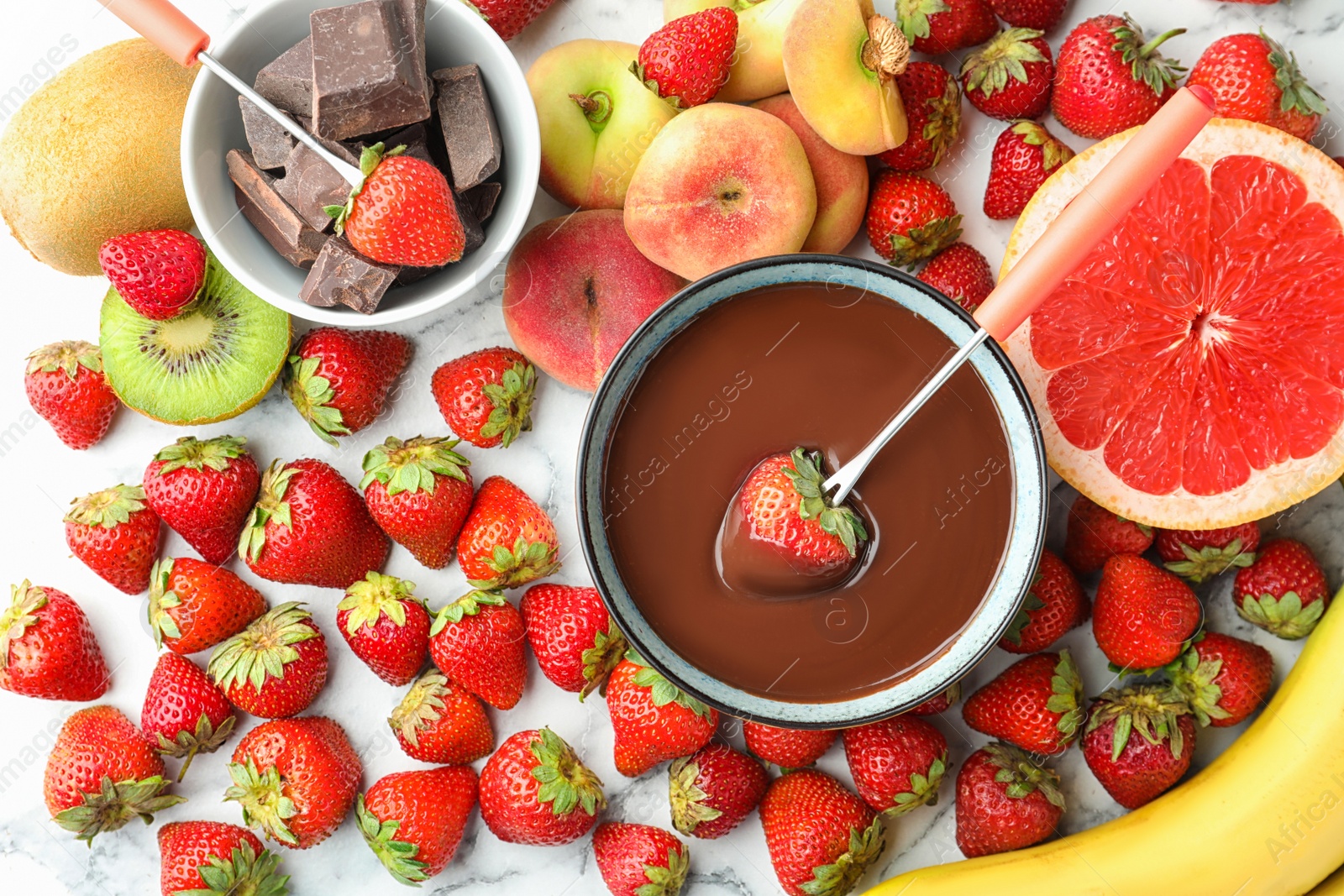 Photo of Fondue fork with strawberry in bowl of melted chocolate surrounded by other fruits on white marble table, flat lay