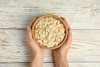 Photo of Young woman with bowl of raw pumpkin seeds at white wooden table, top view