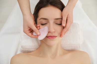 Photo of Young woman receiving facial massage with gua sha tool in beauty salon, top view
