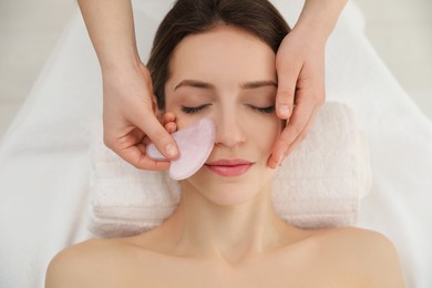 Young woman receiving facial massage with gua sha tool in beauty salon, top view