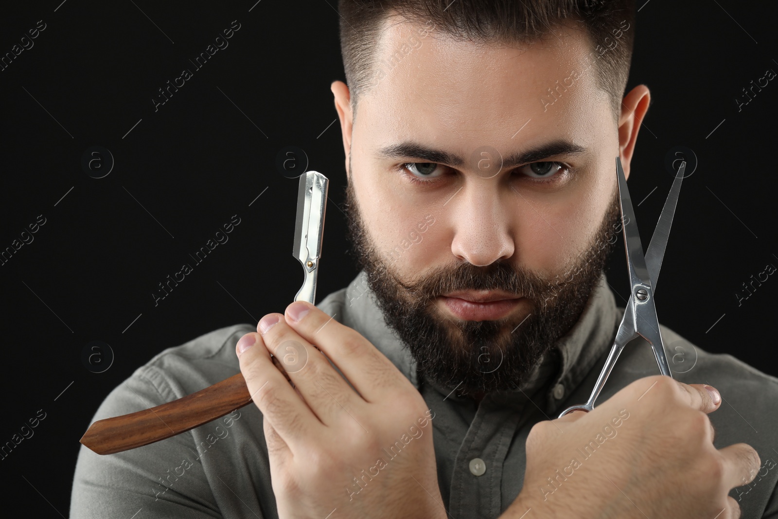 Photo of Handsome young man with mustache holding blade and scissors on black background