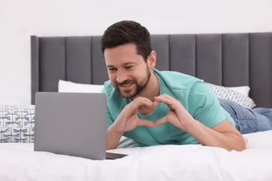 Photo of Happy man making heart with hands during video chat via laptop in bedroom. Long-distance relationship