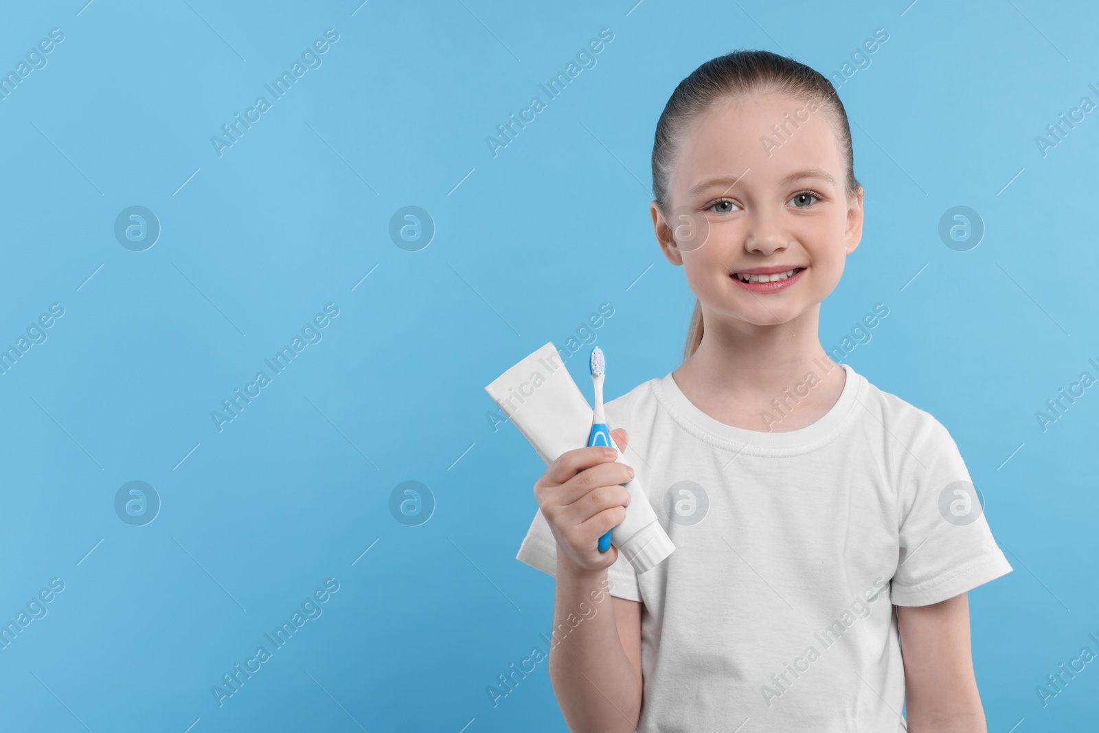 Photo of Happy girl holding toothbrush and tube of toothpaste on light blue background. Space for text