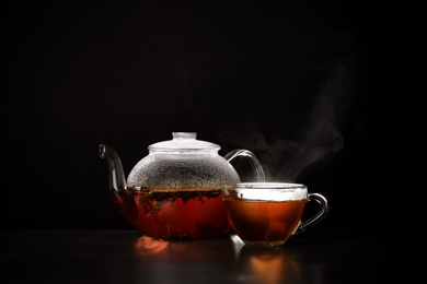 Photo of Glass teapot and cup of hot tea on table against black background, space for text