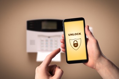 Image of Man operating home alarm system via mobile phone against beige wall with security control panel, closeup. Application interface on device screen