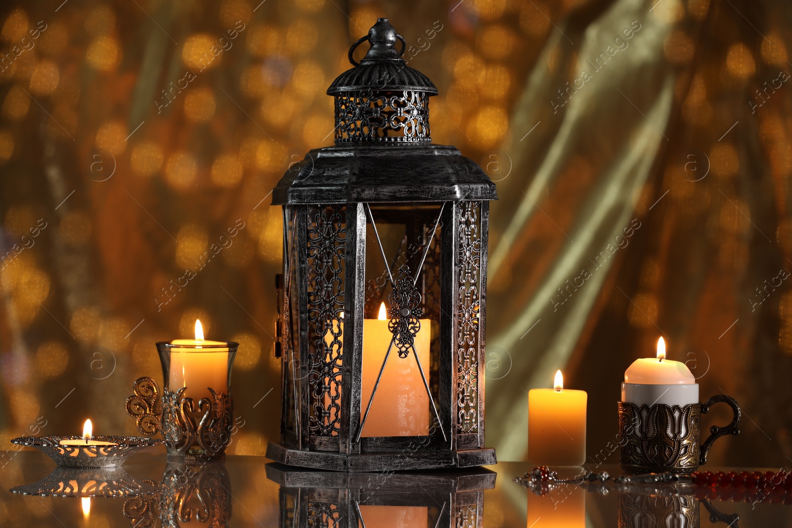 Photo of Arabic lantern and burning candles on mirror surface against blurred lights