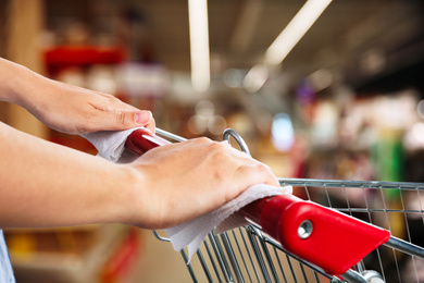 Image of Woman holding shopping cart handle with tissue paper at supermarket, closeup. Preventive measure in public places during coronavirus outbreak