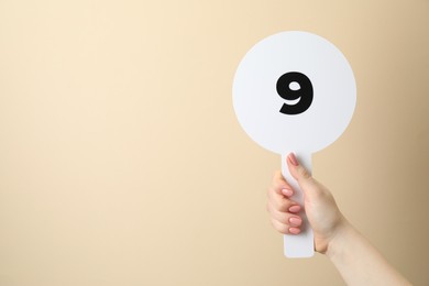 Photo of Woman holding auction paddle with number 9 on beige background, closeup. Space for text