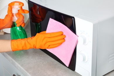 Photo of Woman cleaning microwave oven with rag and detergent in kitchen, closeup