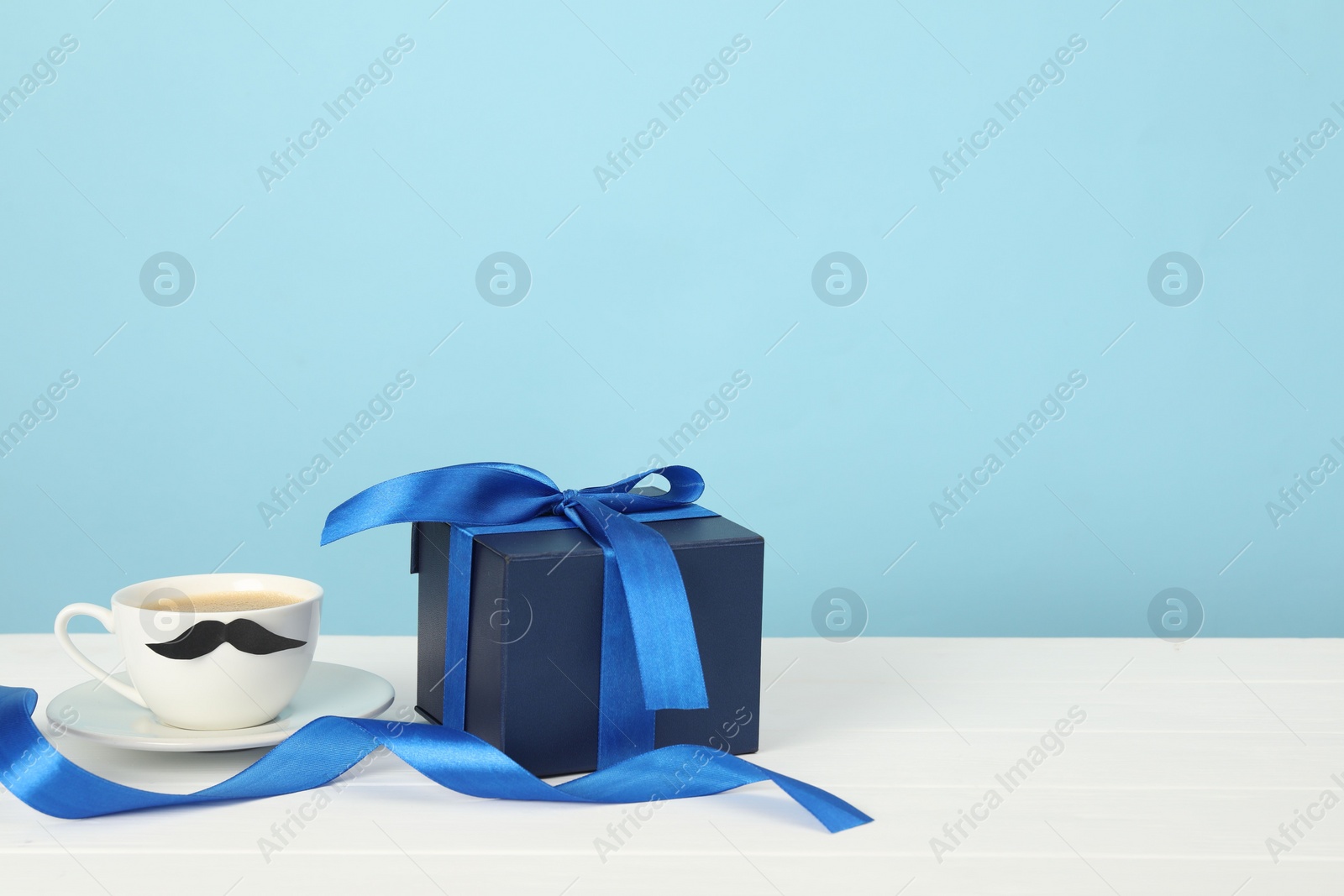 Photo of Happy Father's Day. Cup with moustache and gift box on white wooden table. Space for text