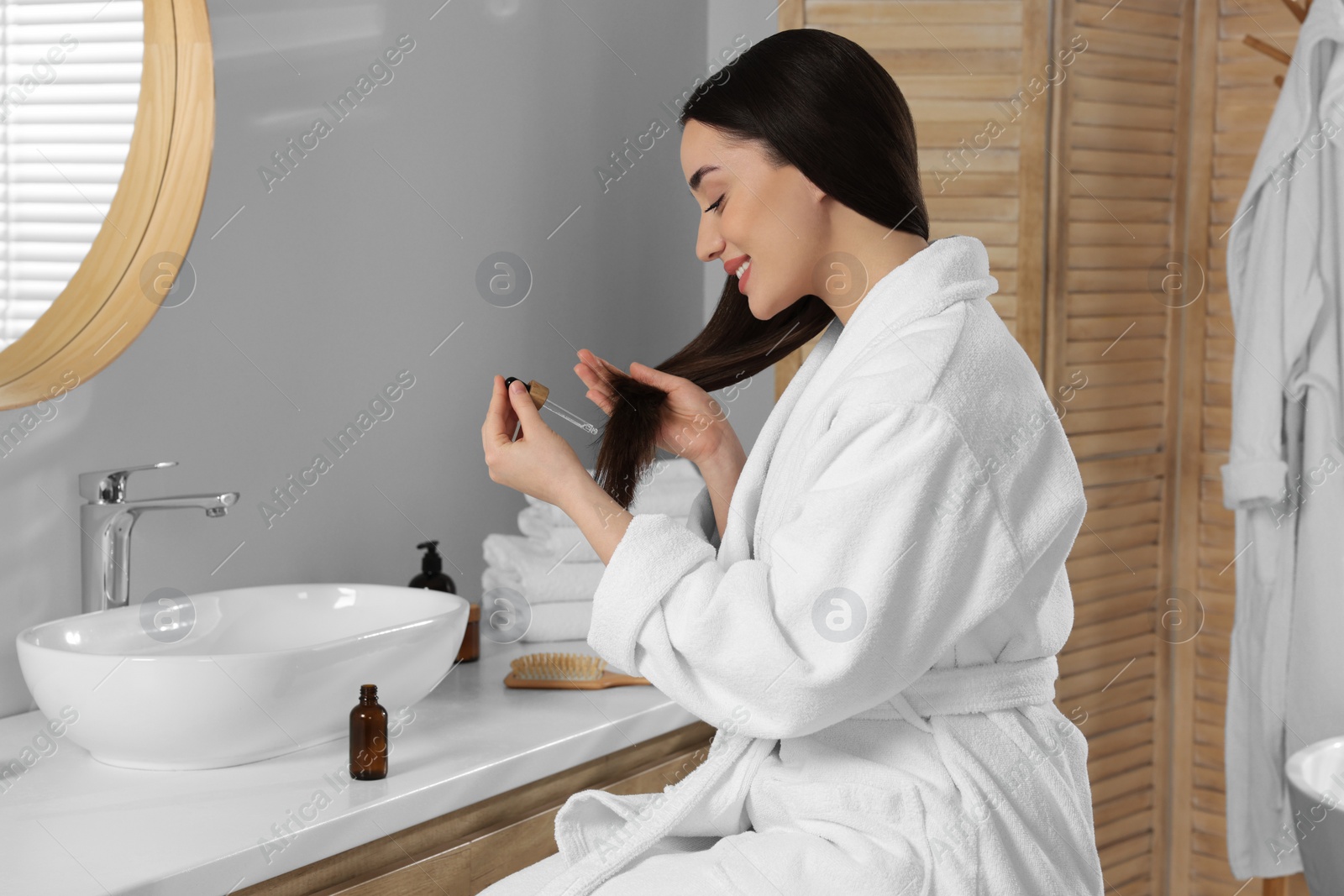 Photo of Happy young woman applying essential oil onto hair in bathroom