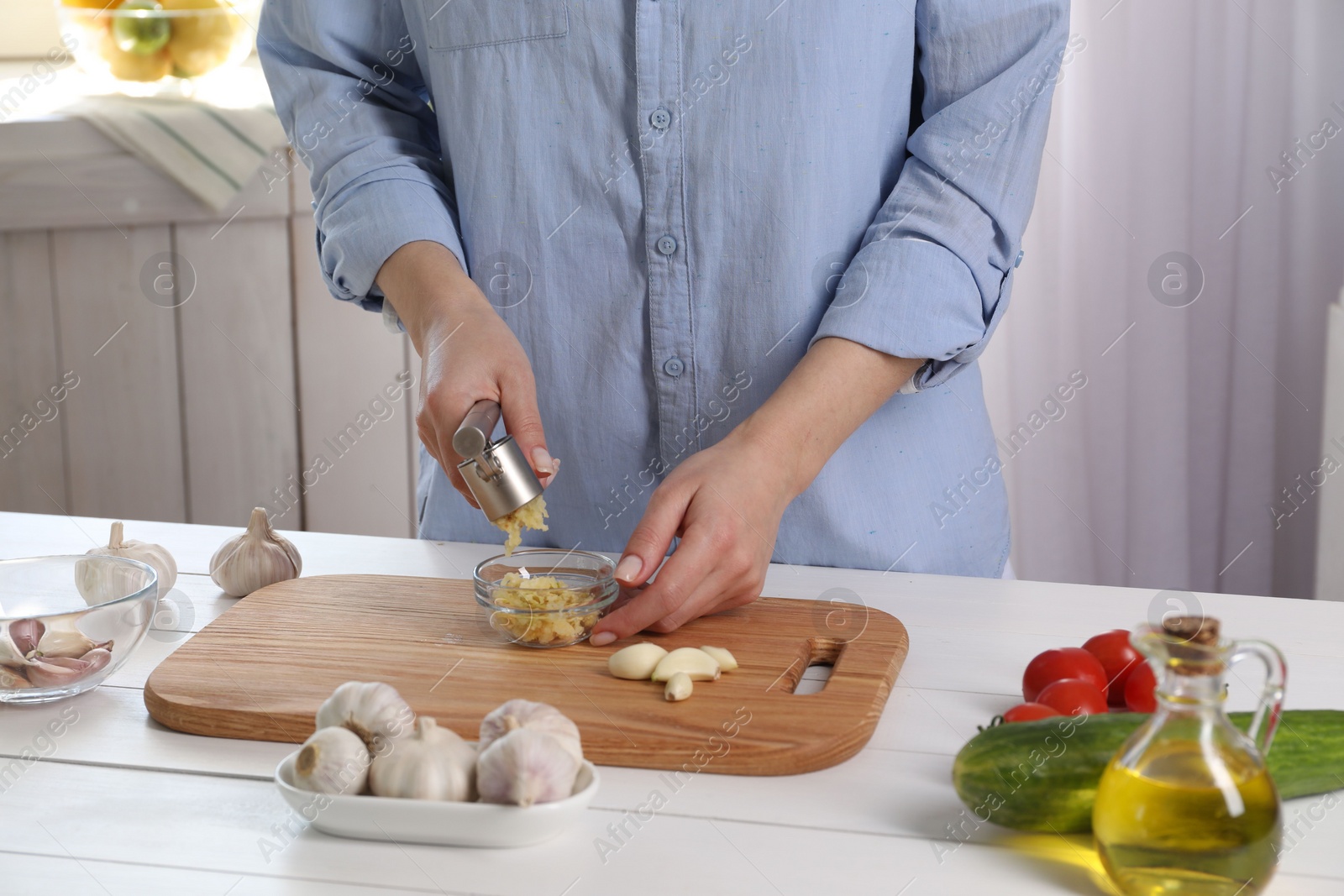 Photo of Woman squeezing garlic with press at white wooden table in kitchen, closeup