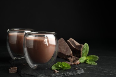 Photo of Glasses of delicious hot chocolate, chunks and fresh mint on black table. Space for text