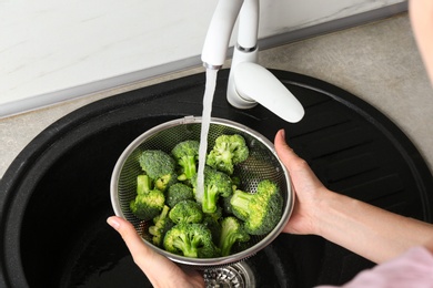 Woman washing fresh green broccoli in metal colander under tap water, closeup view