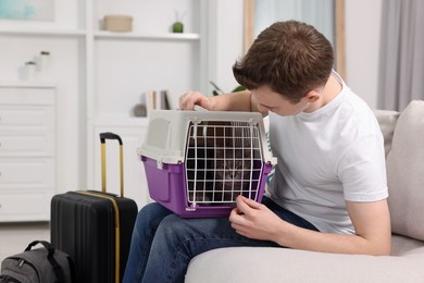 Photo of Travel with pet. Man holding carrier with cute cat on sofa at home
