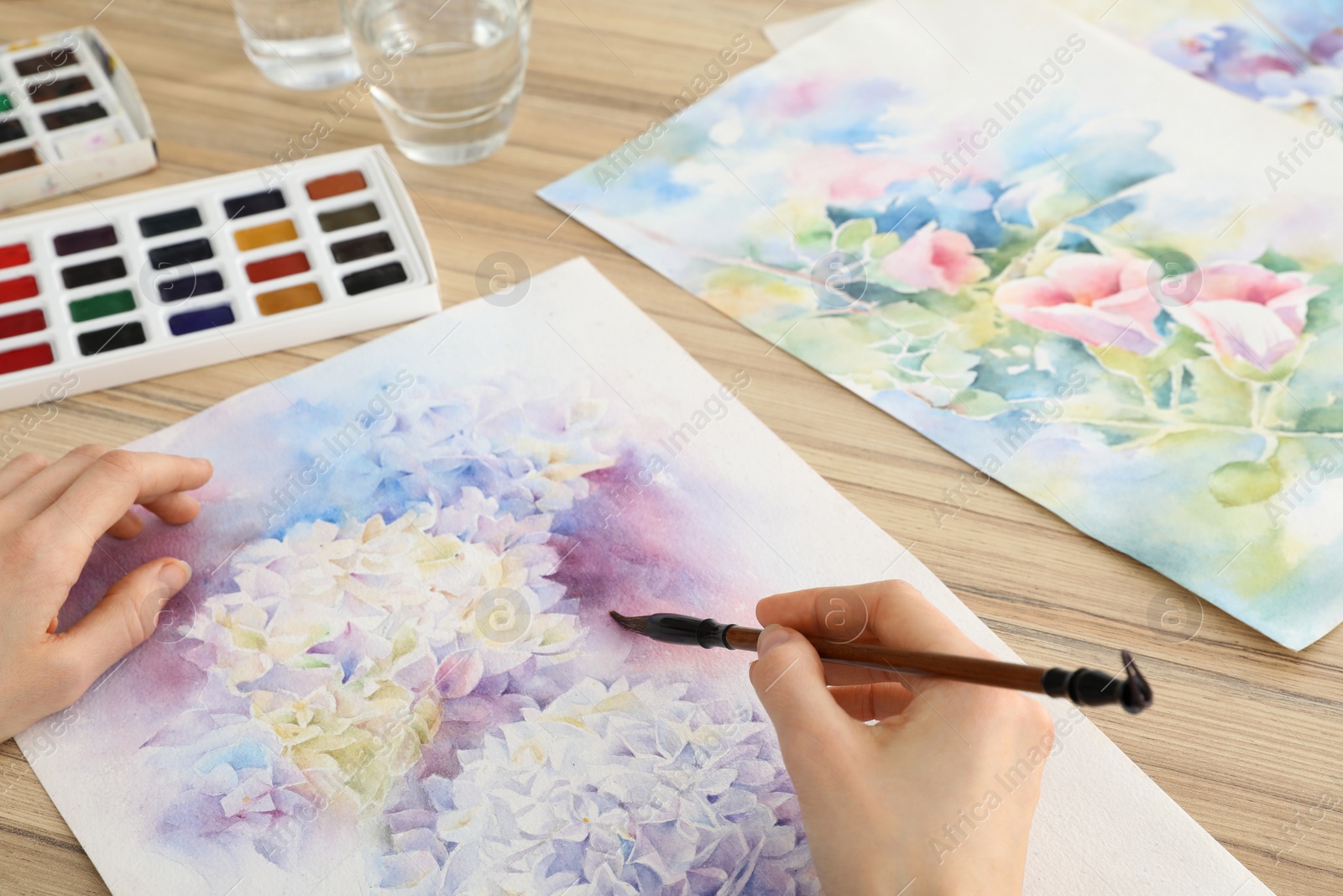 Photo of Woman painting flowers with watercolor at wooden table, closeup. Creative artwork