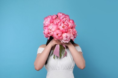 Young woman covering her face with bouquet of pink peonies on light blue background