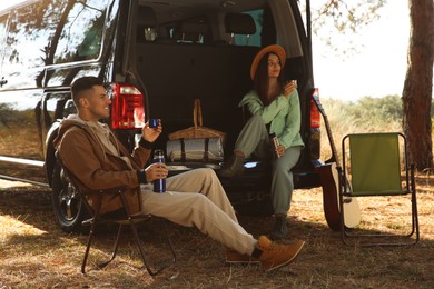 Couple resting near car and enjoying hot drink at camping site